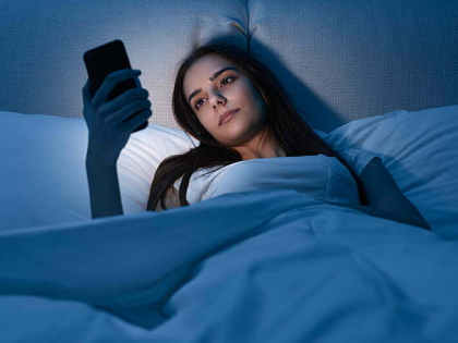 Young woman watching video on cellphone while lying on bed in dark bedroom at night