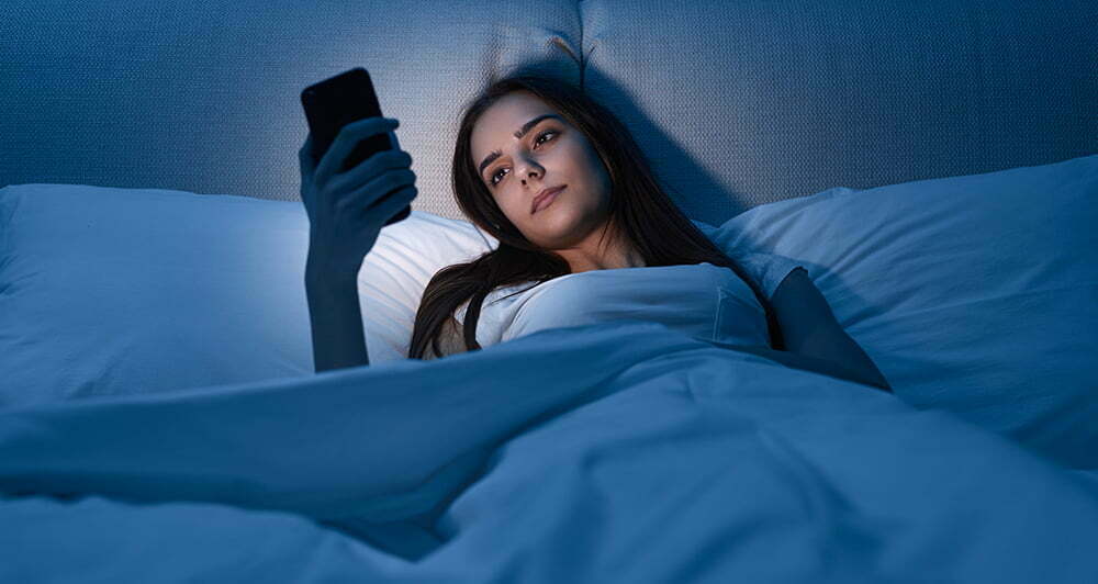 Young woman watching video on cellphone while lying on bed in dark bedroom at night