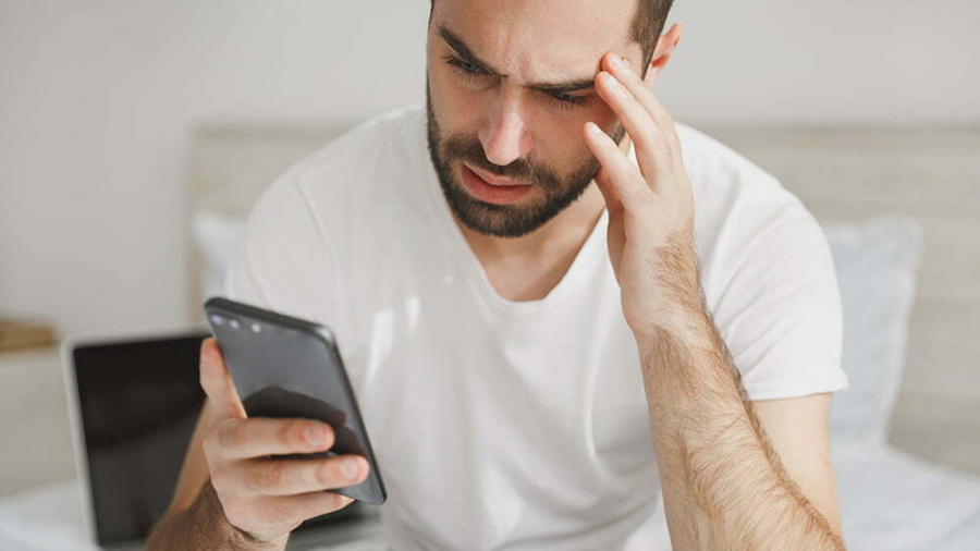 Young handsome bearded man sitting in bed with white sheet pillow blanket in bedroom at home. Male spending time in room using mobile phone. Rest relax good mood lifestyle concept. Mock up copy space