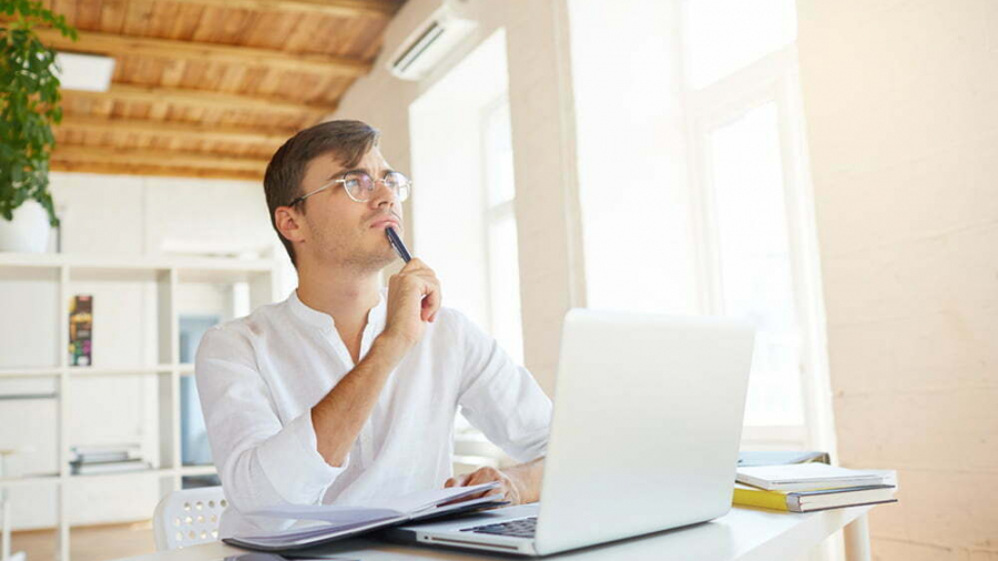 Indoor shot of thoughtful concentrated young businessman wears white shirt and spectacles with laptop and smartphone thinking and writing in notebook at the table in office