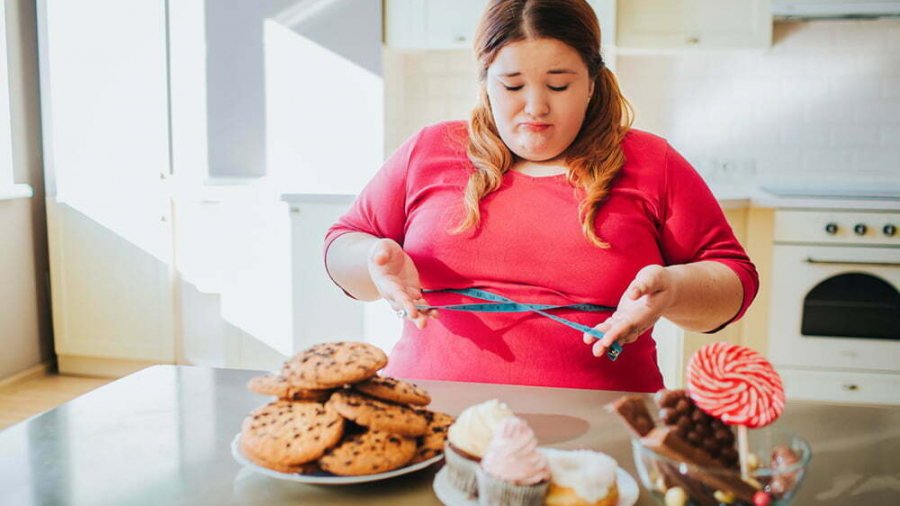 Fat young woman in kitchen sitting and eating sweet food. Confused serious plus size model look on sweets. Body positive. Soft tape measure around waist